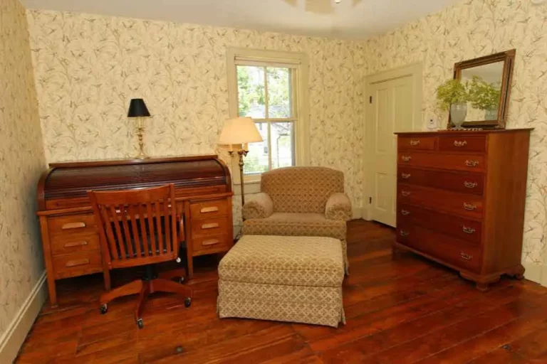 A room with a patterned armchair, wooden furniture, a floor lamp, and a window.