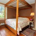 Cozy bedroom with four-poster bed, floral bedding, side table with lamp, and a small rug on hardwood floor.