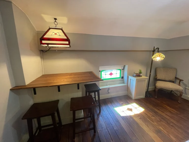 A room with a wooden desk, chairs, a hanging lamp, and a window with blinds.